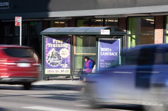 A woman waits for a bus in a shelter at Lincoln at 9th St. as cars rush past.