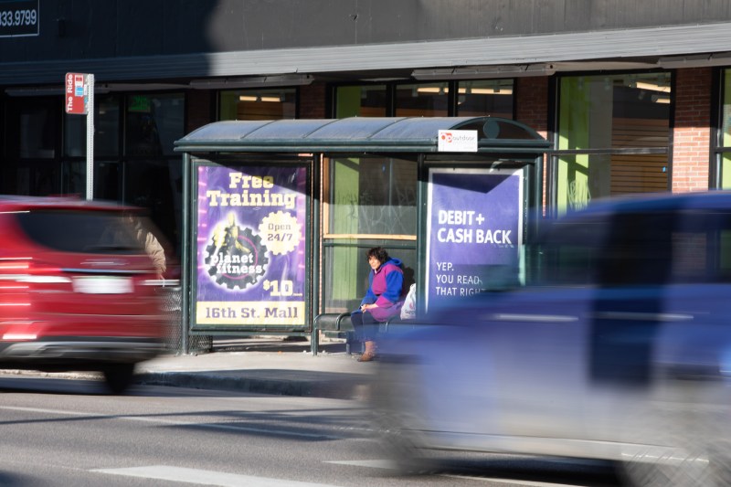 A woman waits for a bus in a shelter at Lincoln at 9th St. as cars rush past.